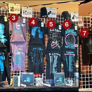 May include: A display of merchandise featuring black hoodies, t-shirts, and hats with various graphic prints and text including Phoebe Bridgers and Lost Weekend. The items are arranged on a wire grid wall with price tags for each product.