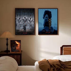 Puede incluir: Tres pósteres de película enmarcados titulados The Odyssey con la fecha 07.17.26. Dos pósteres grandes cuelgan en la pared, uno muestra guerreros con armadura en un bosque y el otro un casco. Un póster más pequeño está sobre una mesa de madera.