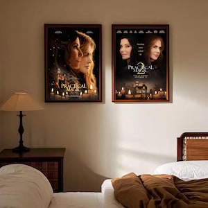 May include: Two framed movie posters for Practical Magic hanging side by side on a wall. Each poster features portraits of two women, glowing candles, and the text Practical Magic. The posters are displayed above a bed and a bedside lamp.
