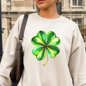 May include: A cream-colored sweatshirt featuring a vibrant, watercolor-style four-leaf clover design. The clover is rendered in shades of green and yellow, with a detailed, botanical illustration. The sweatshirt has a classic crew neck and long sleeves.