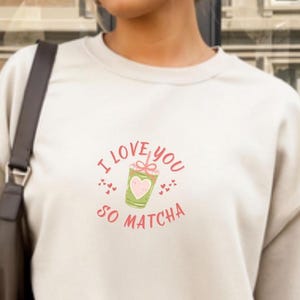 May include: Cream-colored sweatshirt with a graphic that says "I love you so matcha." The design features a green matcha drink with a pink heart and bow, surrounded by pink hearts and text.