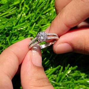 Peut inclure: Une bague argentée dotée d'une pierre centrale ronde transparente montée sur griffes, avec un anneau à double branche orné de petites pierres transparentes serties pavé sur les deux côtés, offrant un aspect brillant et réfléchissant.