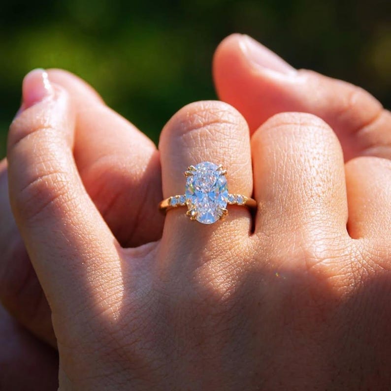 May include: A gold ring with a large, oval-shaped diamond. The ring has smaller stones set into the band. The ring is shown against a blurred green background.