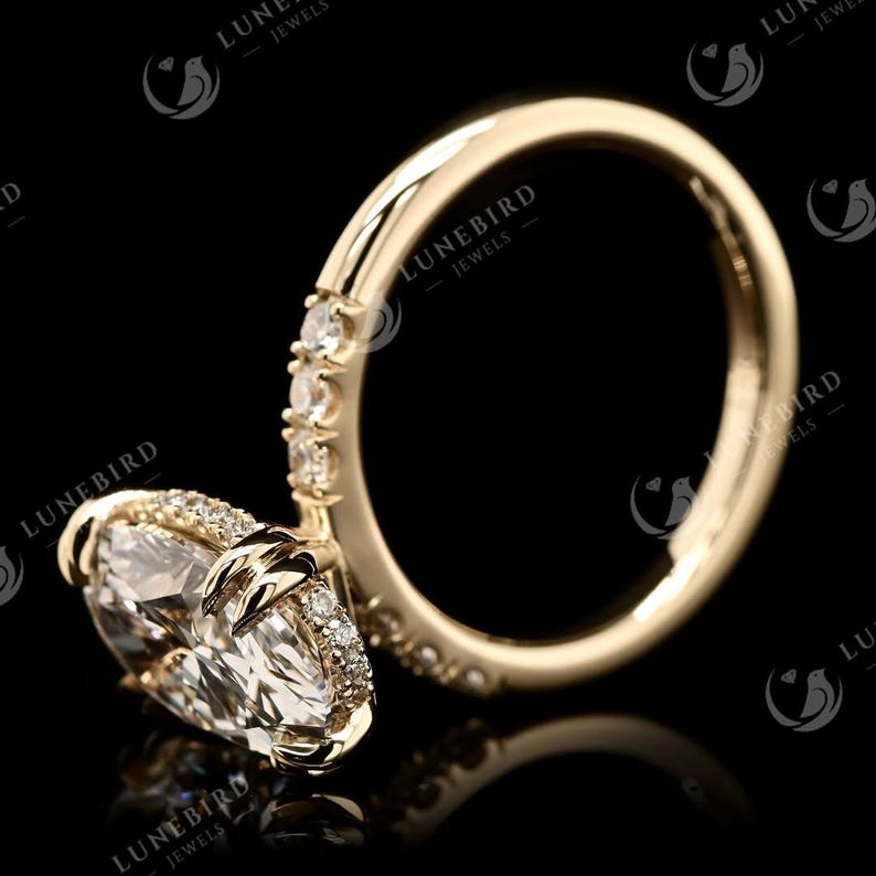 May include: A gold engagement ring featuring a large, oval-cut diamond. The band is embellished with smaller diamonds. The ring is set against a black backdrop, accentuating its sparkle. The ring is from LuneBird Jewels.