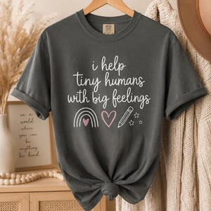 Puede incluir: Una camiseta de manga corta gris carbón con texto serigrafiado en blanco y rosa claro que dice: "i help tiny humans with big feelings". El diseño incluye un arcoíris de líneas simples, un corazón, un lápiz y tres estrellas pequeñas.