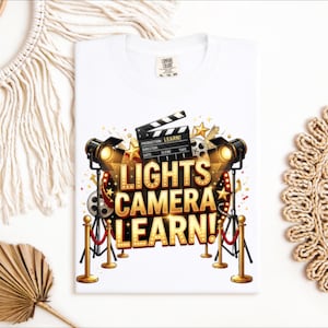 May include: White t-shirt with a colorful graphic design featuring film-related elements. The design includes spotlights, a film clapperboard, and the text "LIGHTS CAMERA LEARN!" in gold lettering. The shirt is a Comfort Colors brand.