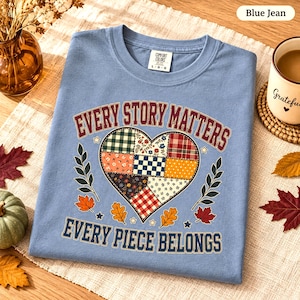 Peut inclure: Un t-shirt bleu jean avec un motif central en forme de cœur composé de divers motifs en patchwork. Le texte "EVERY STORY MATTERS" et "EVERY PIECE BELONGS" est imprimé en rouge foncé et bleu, entouré de petites icônes de feuilles et d'étoiles.