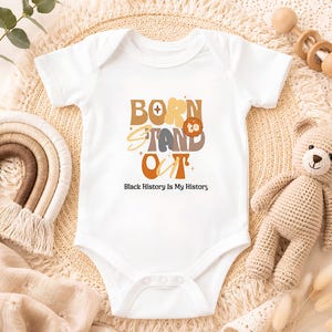 May include: A white short-sleeved baby bodysuit featuring the text 'BORN TO STAND OUT' in a multi-coloured, layered, retro-style font, with the additional text 'Black History Is My History' printed in a simple black sans-serif typeface below.