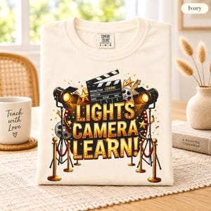 May include: An ivory t-shirt featuring a graphic print with the text LIGHTS CAMERA LEARN! in a marquee style. The design includes a film clapperboard, two studio spotlights, film reels, gold stars, and red velvet ropes on gold stanchions.
