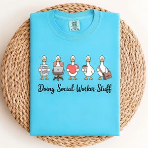 May include: A bright blue t-shirt featuring a graphic of five white ducks in a row. The ducks are holding a document, sitting at a laptop, holding a red heart, holding a coffee cup, and carrying a brown bag. The text reads Doing Social Worker Stuff.