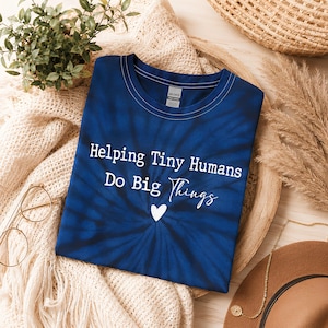 May include: A dark blue tie-dye t-shirt featuring the white text "Helping Tiny Humans Do Big Things" printed in a mix of serif and script fonts, centred above a small white heart icon. The shirt has a crew neckline and a Gildan brand label.