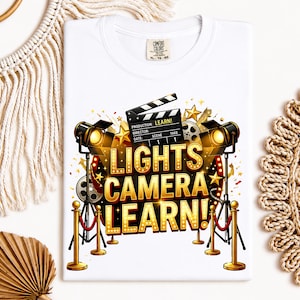 May include: White t-shirt with a graphic design featuring film production elements. The design includes a film clapboard, spotlights, and the text "LIGHTS CAMERA LEARN!" in gold lettering. The shirt has a Comfort Colors tag.