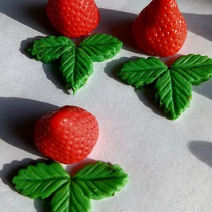 Op de afbeelding: Drie rode aardbeivormige decoraties met getextureerde oppervlakken en groene bladaccenten. De aardbeien zijn gerangschikt met hun bladeren, waardoor een decoratief arrangement ontstaat. De afbeelding is goed verlicht en benadrukt de details.