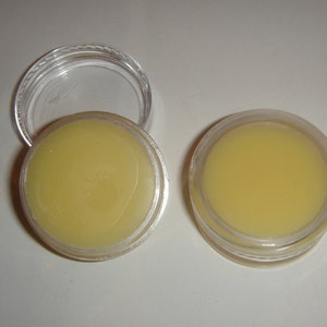 Puede incluir: Dos pequeños recipientes de plástico transparente con tapas, cada uno lleno de un bálsamo o ungüento amarillo.