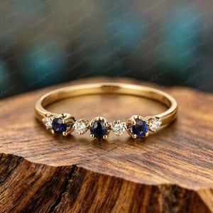 May include: A gold ring featuring a series of alternating dark blue and clear gemstones. The ring's band is a thin, polished gold, and the gemstones are set in delicate, ornate settings. The ring is displayed on a textured wooden surface.