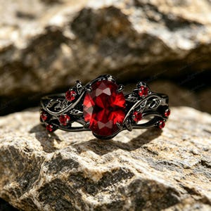 Könnte beinhalten: Ein schwarzer Ring mit einem großen, oval geschliffenen, roten Edelstein als Mittelpunkt. Der Ring hat ein florales Design mit kleineren roten Edelsteinen, vor einem strukturierten, grauen Felsenhintergrund.