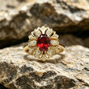 May include: A gold ring with a central oval-cut red gemstone, encircled by a floral arrangement of smaller, clear gemstones. The ring is displayed on a textured, light brown stone surface.