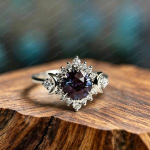 Op de afbeelding: Een zilveren ring met een grote, gefacetteerde, donkerpaarse edelsteen, omringd door kleine, heldere accentstenen. De ring heeft een uniek ontwerp met halve maanvormen aan weerszijden van de centrale steen, op hout.
