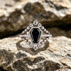 May include: A silver-toned ring featuring a central black coffin-shaped gemstone surrounded by a halo of small round clear stones. The band is split into two rows, each adorned with a series of round clear stones that frame the central setting.