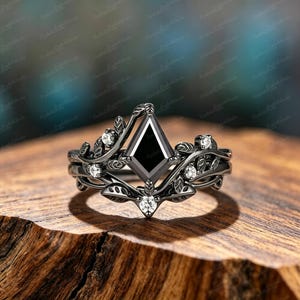 May include: A black metal ring featuring a central kite-shaped black stone. The band is designed with intricate vine and leaf motifs that wrap around the finger, accented by five small, round, clear white stones set within the metal foliage.