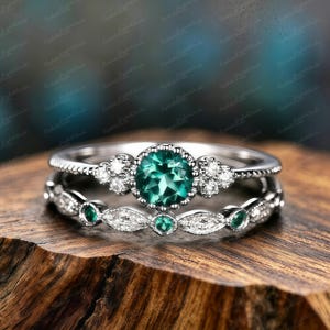 Puede incluir: Un juego de anillos de dos piezas con una banda plateada con una gran piedra preciosa verde esmeralda redonda en el centro. El anillo superior tiene piedras preciosas blancas más pequeñas. El anillo inferior tiene piedras preciosas verdes esmeralda y blancas.
