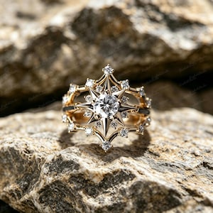 May include: A gold ring with a starburst design, featuring a large, round, clear gemstone at the centre. The ring is adorned with smaller, clear gemstones and set against a textured, grey rock background.
