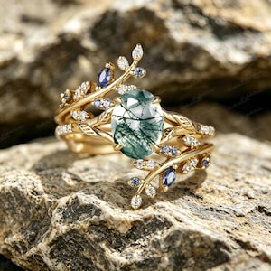 Può includere: Un anello in oro con una pietra centrale ovale in agata muschiata con inclusioni verde scuro. La fascia è decorata con due motivi a ramo impreziositi da piccole pietre bianche trasparenti e pietre blu taglio marquise sulle foglie.