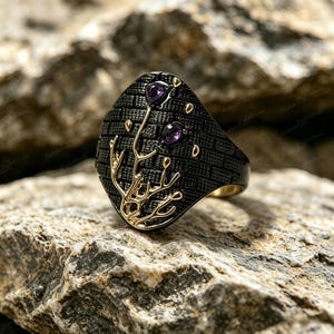 May include: Black and gold ring with a unique design. The ring features a black band with a textured pattern and gold accents. Two purple gemstones are set on the ring, along with a gold branch design.