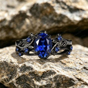 Peut inclure: Une bague de fiançailles noire avec une grande pierre précieuse ovale bleu foncé en son centre. La bague présente un motif floral avec de plus petites pierres bleues et de délicats accents de feuilles. La bague est posée sur un fond texturé de tons neutres.