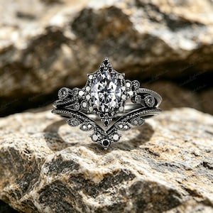Könnte beinhalten: Ein schwarzes Verlobungsring-Set mit einem großen, oval geschliffenen, klaren Edelstein als Mittelpunkt. Der Ring ist von kleineren, runden, schwarzen Edelsteinen und aufwendigen Filigranarbeiten umgeben. Das Band ist ebenfalls schwarz.