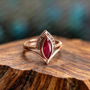 May include: Rose gold ring with a marquise-cut ruby center stone and black accent stones. The ring has a unique, layered design with a V-shaped band. The ruby is a deep red color, and the ring is set on a wooden surface.