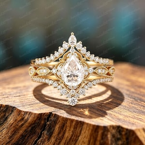 Peut inclure: Une bague en or avec une pierre centrale transparente en forme de poire entourée d'un halo de petites pierres rondes. Le design comprend deux anneaux en forme de chevron ornés de pierres pavées et de détails métalliques complexes.