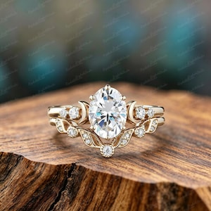 Op de afbeelding: Een gouden verlovingsringset met een ovaalvormige middensteen en een bijpassende band. De ringen hebben kleine accentstenen en een hemels ontwerp met maan- en blad details.
