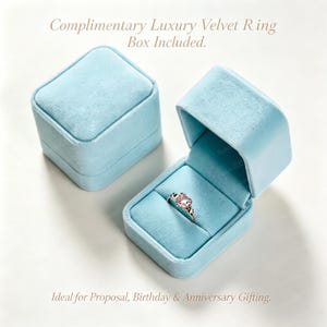 May include: A light blue velvet ring box with a square shape, shown both closed and open. Inside the open box sits a silver ring featuring a heart-shaped pink gemstone. The text "Complimentary Luxury Velvet Ring Box Included" and "Ideal for Proposal, Birthday & Anniversary Gifting" is visible.