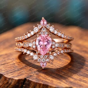 May include: A rose gold ring set featuring a central pear-cut pink gemstone surrounded by smaller clear stones. The design includes a chevron-shaped band adorned with multiple clear stones, creating a layered appearance with pink and clear accents.