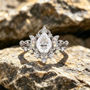 Può includere: Un anello color argento con una pietra centrale trasparente a forma di pera circondata da una montatura in filigrana. La fascia è decorata con piccole opali rotonde e pietre trasparenti disposte in un motivo a mezzaluna.