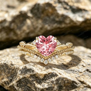 Op de afbeelding: Een gouden verlovingsringset met een hartvormige roze edelsteen en een bijpassende band. De ringen zijn bezet met kleine, heldere edelstenen. De ringen worden gepresenteerd op een getextureerd, lichtbruin oppervlak.