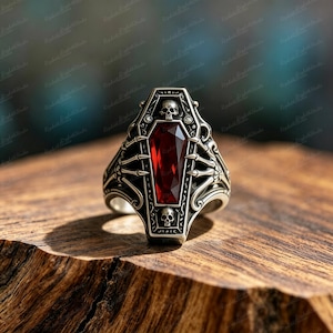 May include: A silver-toned ring featuring a coffin-shaped centerpiece with a faceted deep red gemstone. The band is decorated with skeletal hand motifs reaching toward the center, accented by two small skulls at the top and bottom edges.