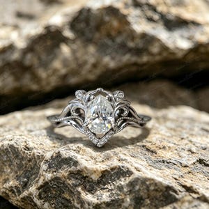 Könnte beinhalten: Ein silberfarbener Ring mit einem zentralen, birnenförmig geschliffenen, klaren Edelstein. Die Ringschiene zeigt filigrane Verzierungen und Schnörkel, akzentuiert durch kleine, runde, klare Steine entlang der Fassung und der Basis des Steins.