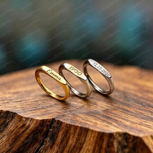 Puede incluir: Tres anillos personalizados con texto grabado. Uno dorado con el nombre "Emma", otro plateado con "1990", y el tercero plateado con la fecha "12.25.2020". Los anillos se exhiben sobre una superficie de madera.