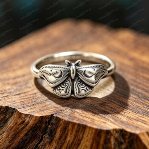 Puede incluir: Un anillo de metal plateado con un diseño detallado de polilla como pieza central. La polilla tiene alas simétricas con patrones grabados, incluyendo formas de media luna y líneas intrincadas, sobre una banda circular lisa y pulida.