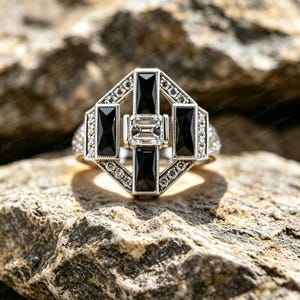 Puede incluir: Un anillo de tono plateado con una piedra central rectangular transparente rodeada por cuatro piedras rectangulares negras alargadas en forma de cruz, con pequeñas piedras redondas transparentes en el marco geométrico exterior.