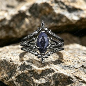 Puede incluir: Un anillo de metal negro con una piedra central de arenisca azul oscuro de corte marquesa con inclusiones brillantes. La banda presenta un marco geométrico puntiagudo adornado con pequeñas piedras negras redondas a lo largo del cuerpo.
