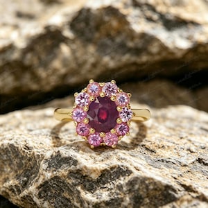 Pode incluir: Um anel de ouro com uma grande pedra central vermelho-escura de corte oval, rodeada por um halo de doze pedras mais pequenas de corte redondo cor-de-rosa claro. O aro é liso e polido, apresentando um design clássico de grupo floral.