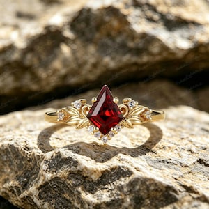 May include: A gold ring with a striking, kite-shaped, deep red gemstone. The ring is embellished with smaller, clear gemstones and leaf-shaped details. The ring is displayed on a textured, natural stone surface.