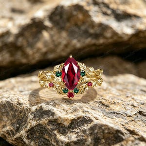 Pode incluir: Um anel dourado com uma grande pedra preciosa vermelha escura com corte marquise como ponto focal. O anel é adornado com pedras preciosas vermelhas e verdes menores e um design detalhado de folhas. O anel está sobre uma superfície de pedra texturizada marrom claro.