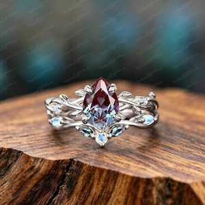 Può includere: Un anello color argento con una pietra centrale a goccia che cambia colore, passando dal rosso intenso al blu-verde. La fascia è decorata con intricati motivi in metallo a forma di viticci e foglie, arricchiti da piccole pietre di luna rotonde.