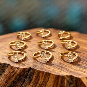 Könnte beinhalten: Eine Sammlung von neun goldfarbenen Ringen, die jeweils mit einem Buchstaben des Alphabets und kleinen klaren Steinen besetzt sind. Die Ringe sind in drei Reihen auf einer strukturierten Holzoberfläche angeordnet und zeigen die Buchstaben M, Y, Y, R, V, E, U, W und Q.