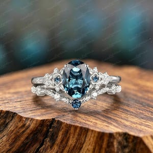Può includere: Un set di anelli color argento con una grande pietra centrale blu scuro taglio ovale, accentuata da pietre blu e trasparenti più piccole incastonate in una fascia metallica floreale con dettagli a forma di foglia sull'anello e sulla fede abbinata.