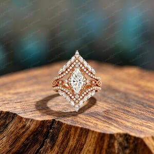 Könnte beinhalten: Ein Verlobungsring-Set aus Roségold mit einem Mittelstein im Marquise-Schliff und Diamantakzenten. Der Ring hat ein einzigartiges, verziertes Design mit einem passenden Band, das auf einer Holzoberfläche liegt. Ein Symbol der Liebe.